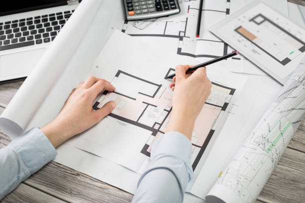 Architecte dessinant un plan de maison sur un bureau avec rouleaux de plans, ordinateur et calculatrice. Conception et modélisation de projet immobilier sur mesure