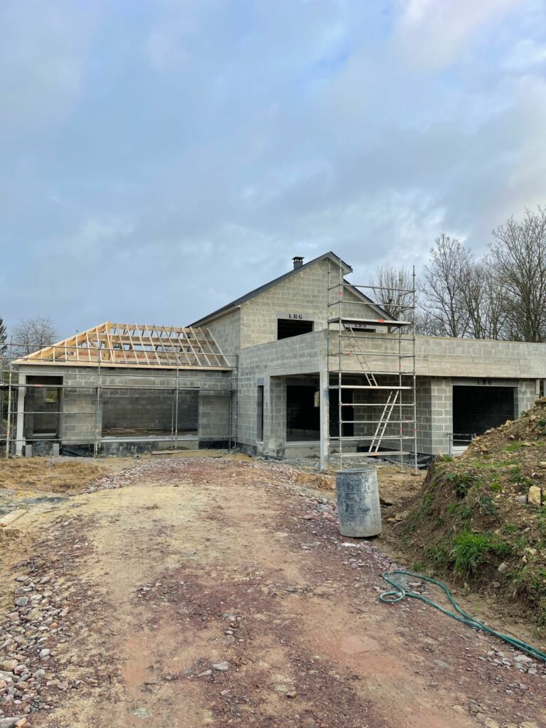 Maison en construction avec murs en parpaings et charpente en cours d’installation. Projet de maison moderne en phase de gros œuvre avec échafaudage et accès chantier.